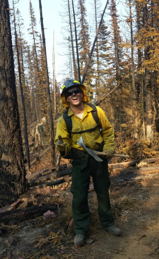 A person in yellow fire gear standing in a forest smiling at the camera.